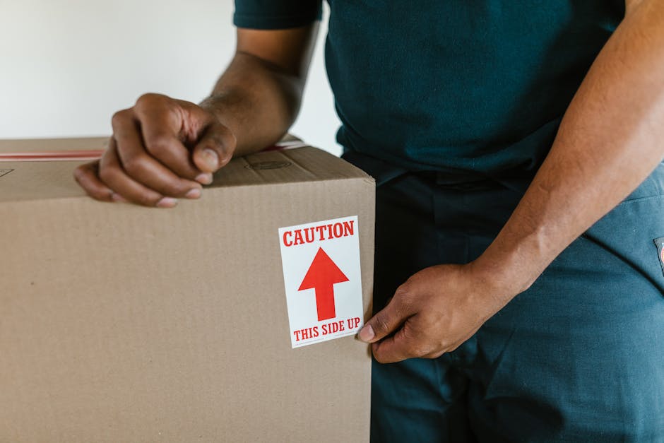 Close-up of a person wearing a dark green or navy blue uniform, sealing a rectangular cardboard box with packing tape during a home relocation process. The box has a white label with red text and an u