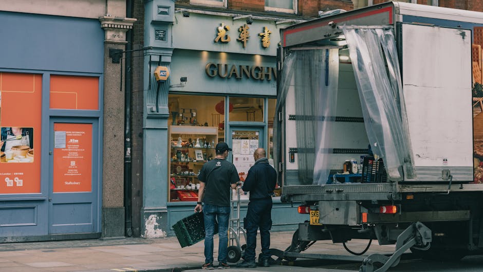 Two men are standing outside a shop named 'GUANGH' on a city street, engaged in a home relocation or furniture transport process. One man is holding a plastic crate, while the other has a hand truck w